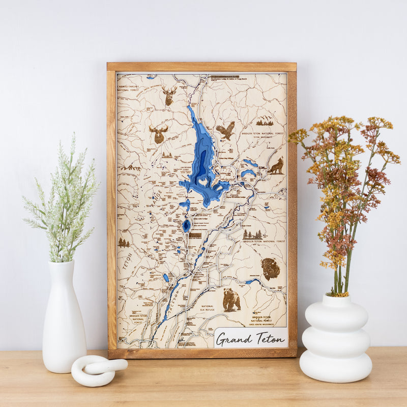 Laser-engraved wooden map of Grand Teton National Park with layered blue inlay and trail details framed and styled on a tabletop with floral vases.