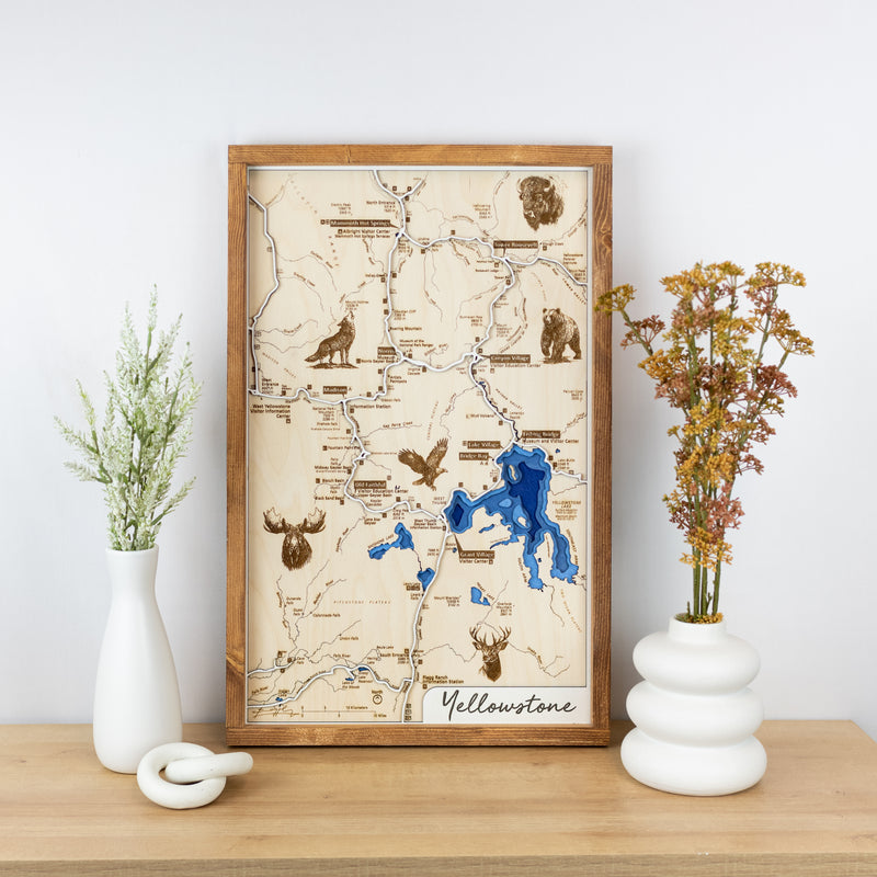Laser-engraved wooden trail map of Yellowstone National Park with blue water layers, animals, and trails resting on a shelf.